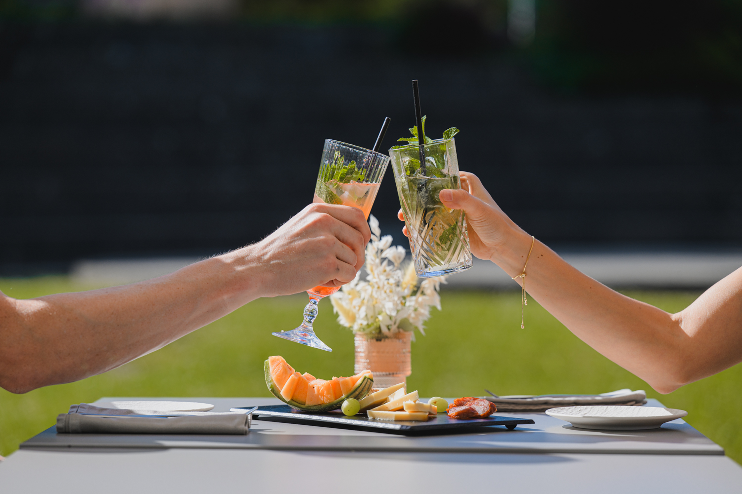 Deux personnes trinquent en terrasse autour d’une planche de fromages et de charcuterie.